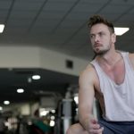 Muscular man with facial hair sitting and looking away in a gym, captured in selective focus.