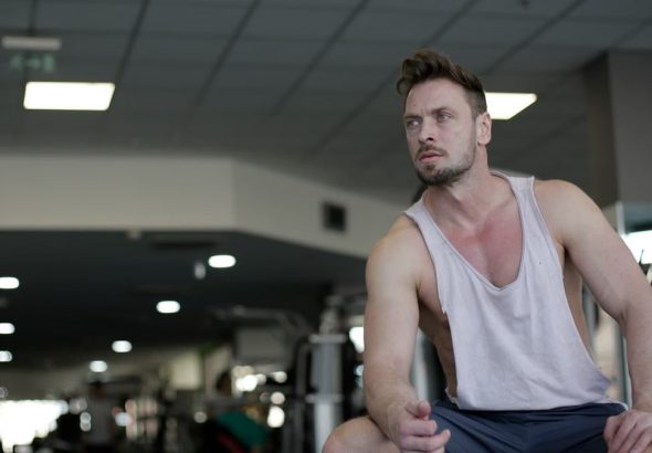 Muscular man with facial hair sitting and looking away in a gym, captured in selective focus.