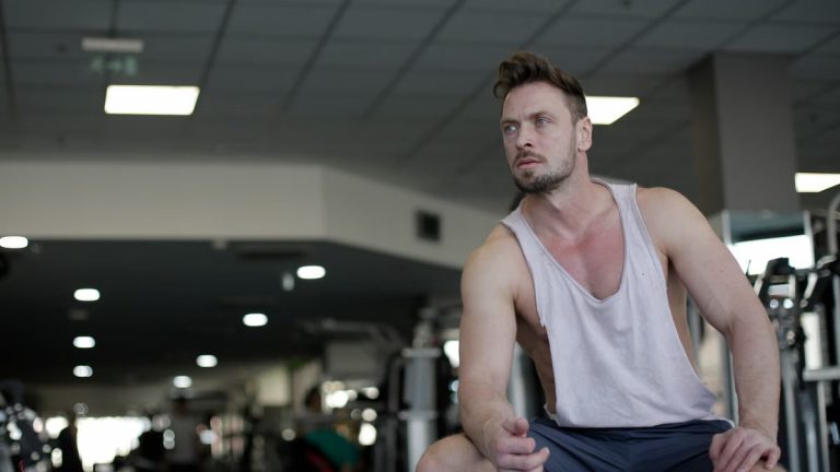 Muscular man with facial hair sitting and looking away in a gym, captured in selective focus.